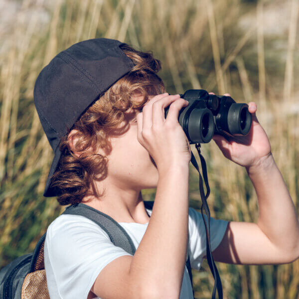 Blond boy with cap and backpack admiring nature through binoculars while on blurred background of tall grass near Pollensa town on sunny day in Mallorca, Spain