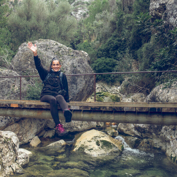 Full length of cheerful young Hispanic female hiker in casual outfit smiling and waving hand while sitting on aged wooden footbridge over river during trekking in Biniaraix ravine